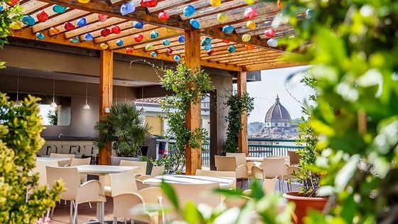 Rooftop cafe with colorful lights, wooden pergola, and greenery, offering a view of a historic dome in the background on a sunny day.