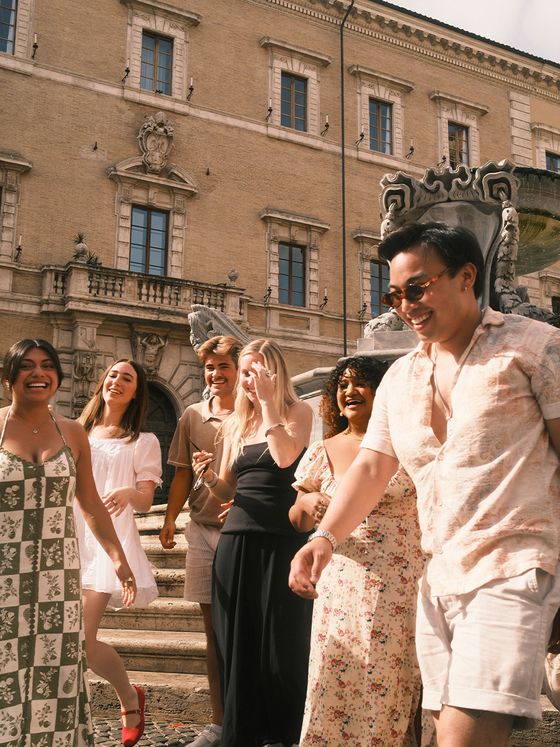A group of six people smiling and walking in front of a historic building with ornate architecture and a stone fountain.