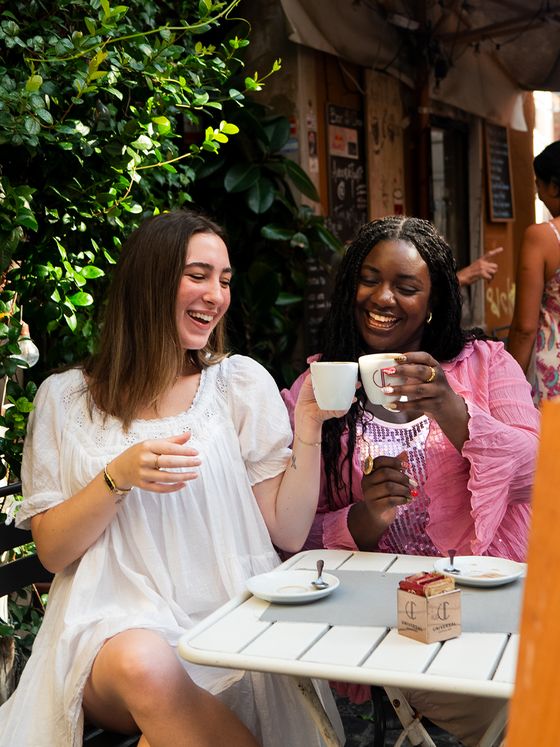 Two women laughing and clinking coffee cups at an outdoor café table, surrounded by greenery and a vibrant street scene.