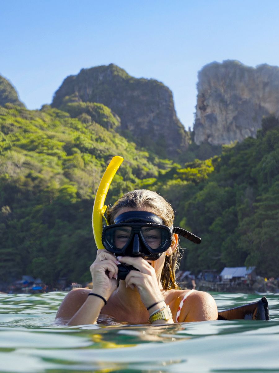 A person wearing snorkeling gear floats in clear water with lush green cliffs and blue sky in the background.