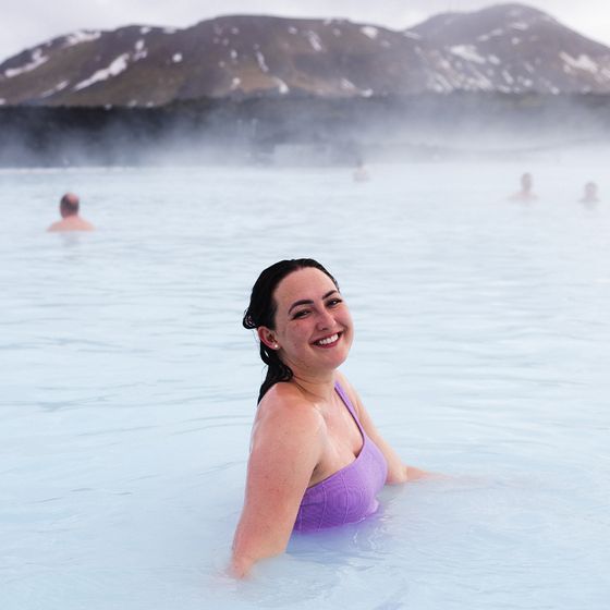 A smiling woman in a purple swimsuit enjoys a geothermal hot spring, with mist and snowy mountains in the background.