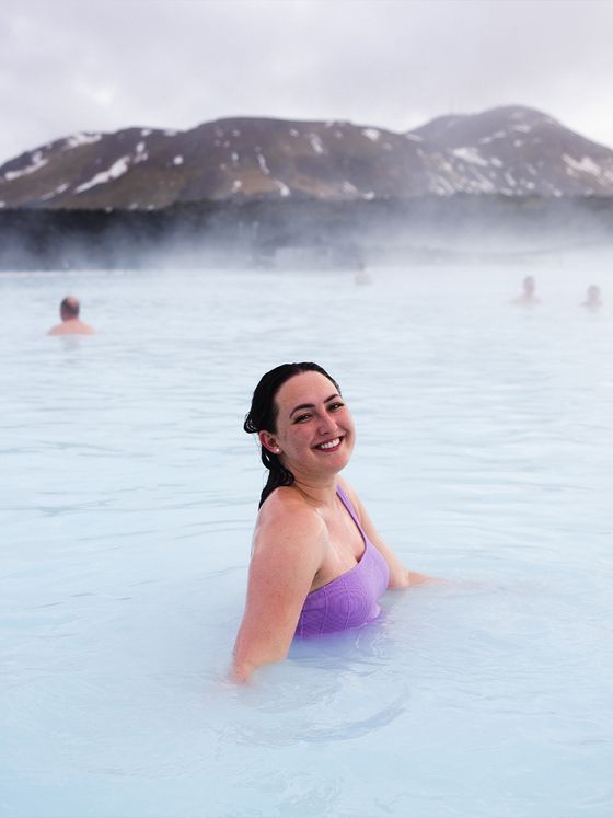 A smiling woman in a purple swimsuit enjoys a geothermal hot spring, with mist and snowy mountains in the background.