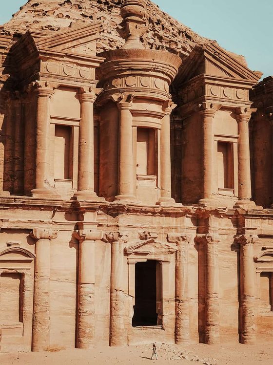 A person stands in front of the ancient stone-carved Al Deir monastery in Petra, Jordan, under a clear blue sky.