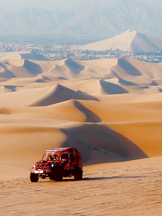 A red off-road vehicle drives across vast desert sand dunes, with distant mountains and a city on the horizon under a clear sky.