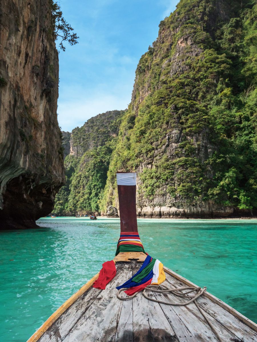 A boat with colorful cloths on its prow navigates clear turquoise waters between lush, towering cliffs under a blue sky.
