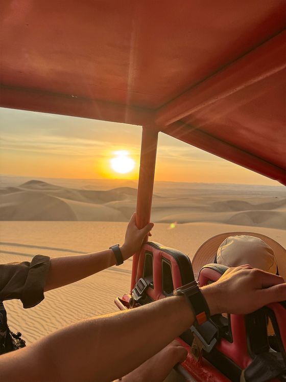 View from a dune buggy over sand dunes at sunset. Two people hold onto the frame, wearing watches. A hat rests on a seat.