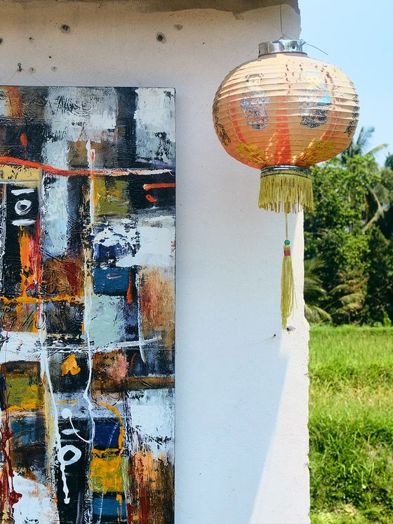 A colorful abstract painting hangs on a white wall beside a traditional paper lantern. Lush greenery is visible in the background.