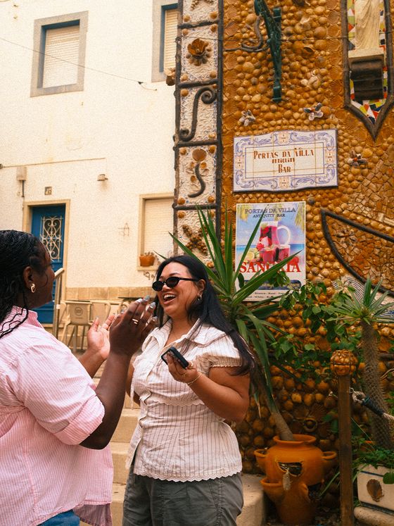 Two people laughing and talking in front of a decorative building façade with a sign that reads "Portal de la Villa Bar."