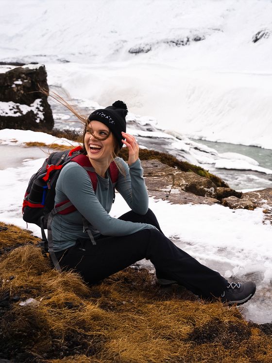 Woman seated on snowy terrain, smiling, with a backpack and beanie, near a river and snow-covered landscape. Hair blowing in the wind.