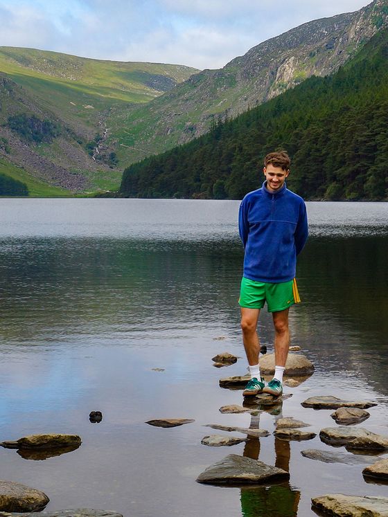 A person in a blue jacket and green shorts standing on rocks at a lake with hills and greenery in the background.