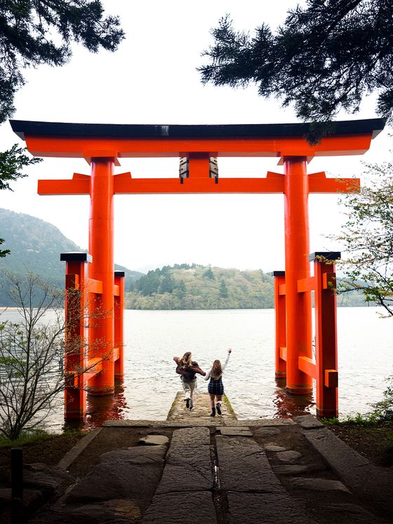 Two people walk towards a large red torii gate by the water, surrounded by trees, with mountains in the background.