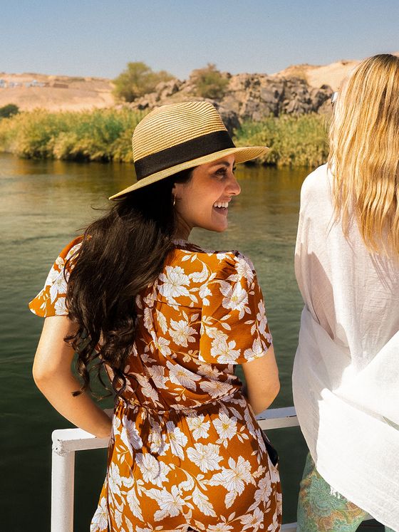 Two women on a boat by a river; one wears a floral dress and hat, smiling, while the other looks away, with scenic greenery in the background.