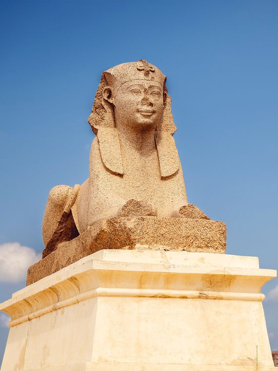Ancient stone sphinx statue on a white pedestal against a clear blue sky, featuring a human face and lion's body.