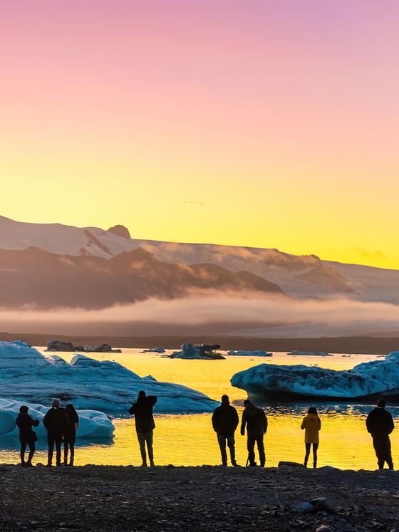 Silhouetted people stand by an icy lake at sunrise or sunset, with vibrant orange and pink skies over snow-capped mountains in the background.