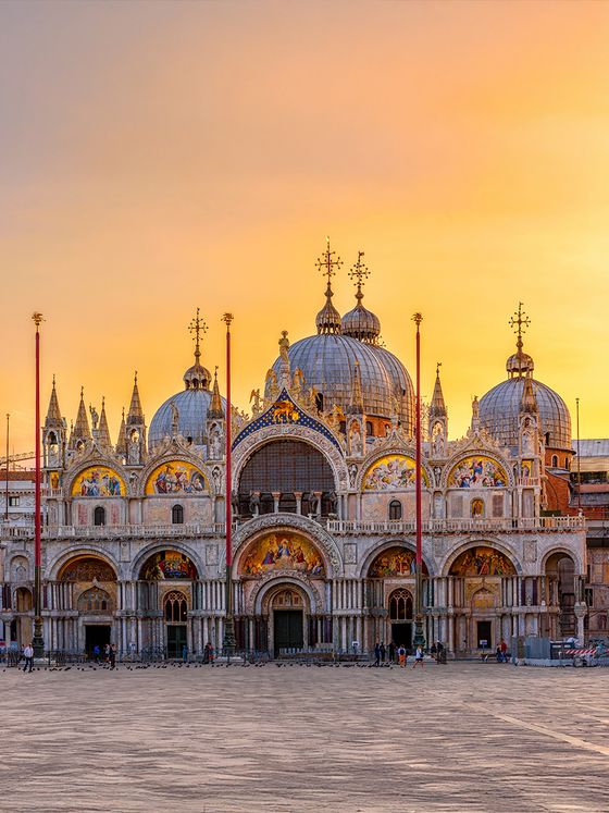 St. Mark's Basilica in Venice at sunset, showcasing its domes and ornate facade against a vibrant orange sky.
