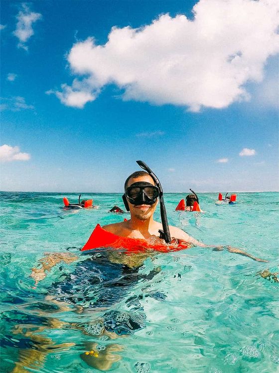 Person snorkeling in clear turquoise water with others in the background, wearing a mask and snorkel, under a partly cloudy sky.