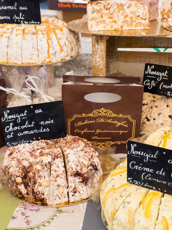 Display of various nougat cakes with handwritten labels, featuring flavors like chocolate almond and lemon. A branded box is visible in the background.