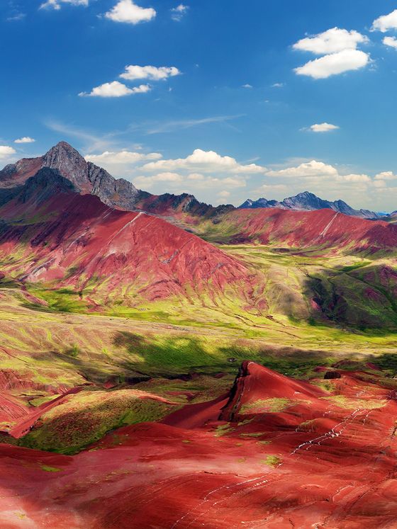 Vibrant landscape of red and green mountains under a blue sky with clouds, featuring rugged peaks and rolling hills.
