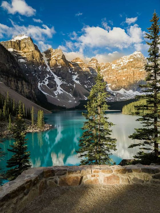 A scenic view of a turquoise lake surrounded by snow-capped mountains and pine trees under a blue sky with clouds.