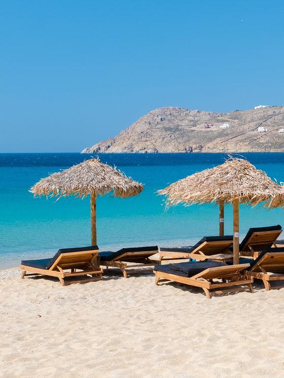 Straw umbrellas and wooden loungers on a sandy beach with clear blue water and distant rocky hills under a clear sky.
