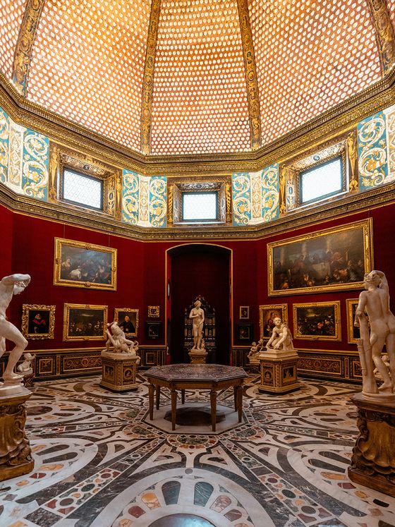 Ornate museum room with a domed ceiling, red walls, classical statues, paintings, and a round table on an intricately patterned floor.