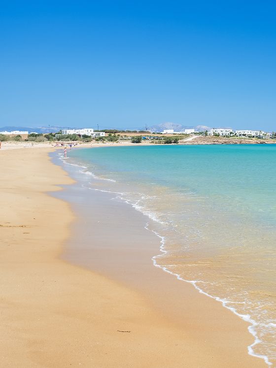 Sandy beach with gentle waves under a clear blue sky; distant shoreline with white buildings and greenery in the background.