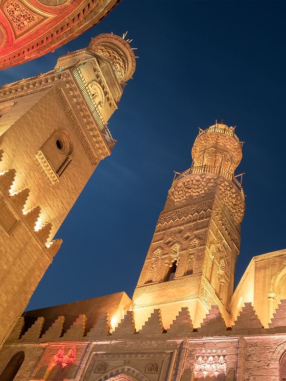 Illuminated historic towers with intricate designs, set against a deep blue evening sky, showcasing Middle Eastern architectural beauty.