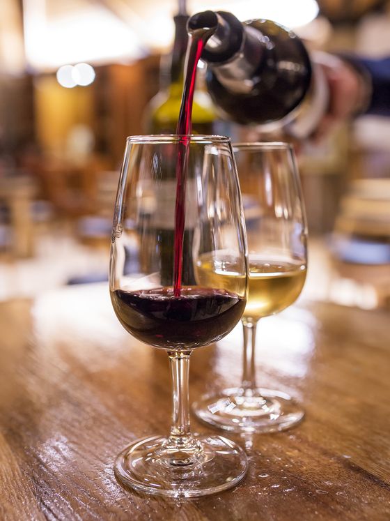 Close-up of red wine being poured into a glass beside a filled glass of white wine on a wooden table in a warmly lit setting.