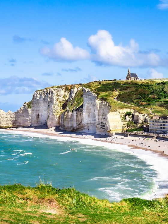 White cliffs of Étretat with a quaint chapel atop. Sandy beach below, turquoise waves, and a clear blue sky dotted with clouds.