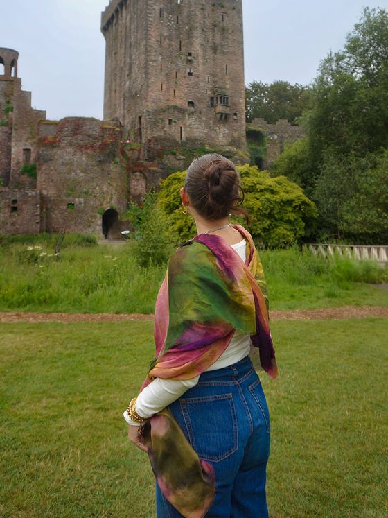 A woman with a colorful scarf looks at a historic stone castle surrounded by greenery on a cloudy day.