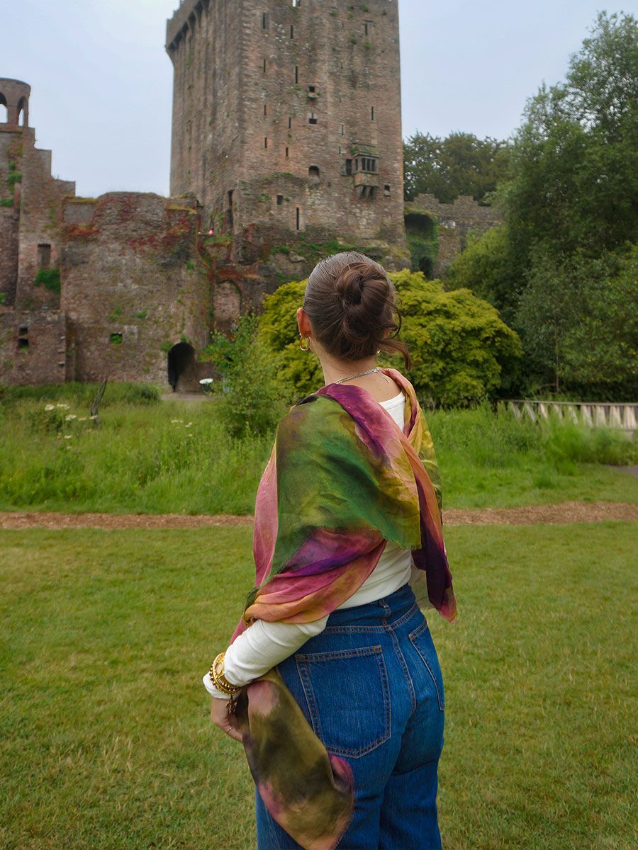 Woman in colorful scarf and jeans stands on grass, facing an ancient stone castle with lush greenery around.