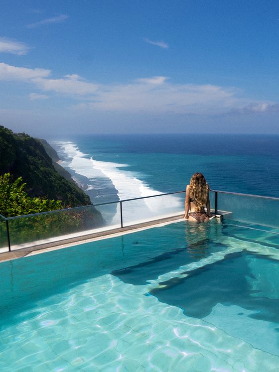 A woman sits at the edge of an infinity pool overlooking a vast ocean and rugged coastline under a clear blue sky.