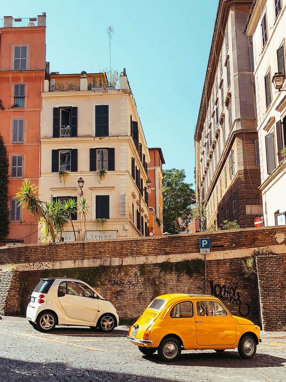 A sunny street scene in Italy with a yellow vintage car and a white compact car parked near colorful, historic buildings.