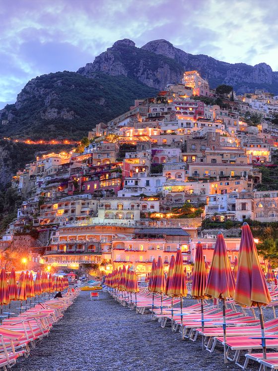 Colorful beach umbrellas line a pebbled shore, with illuminated hillside buildings in Positano under a twilight sky.