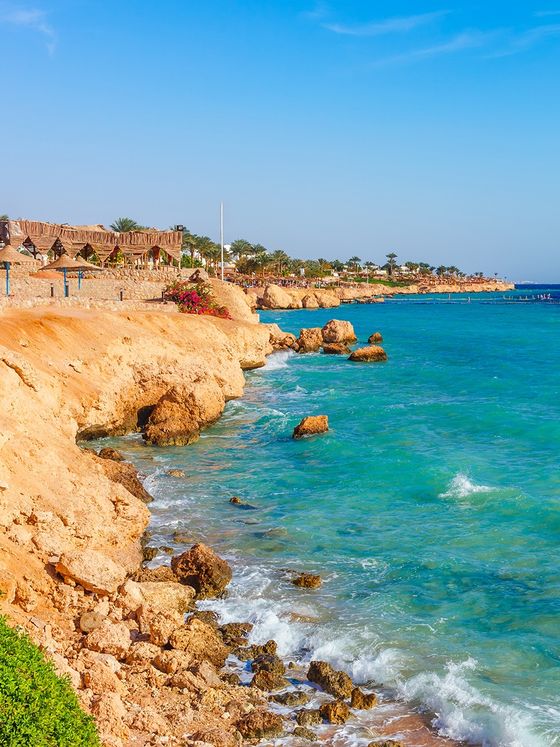 Rocky coastline with turquoise waves hitting the shore under a clear blue sky, lined with thatched-roof huts and palm trees in the background.