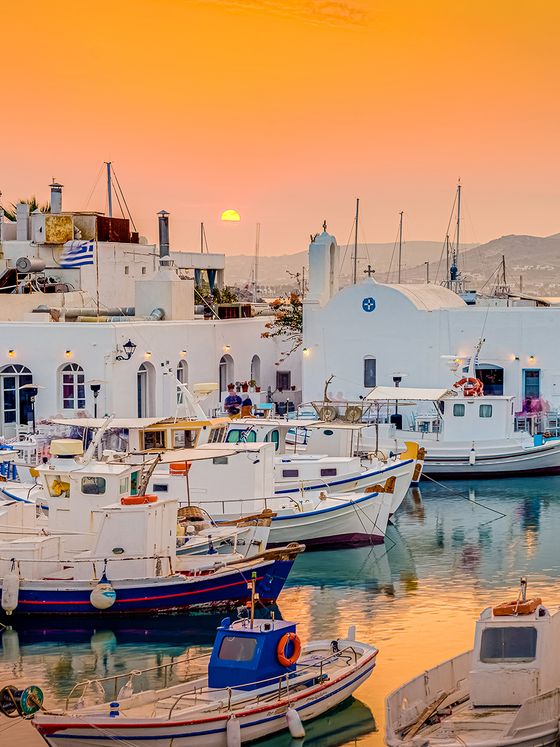 Idyllic harbor at sunset with white buildings and moored boats reflecting on calm water, under a vibrant orange sky. Greek flag on a building.