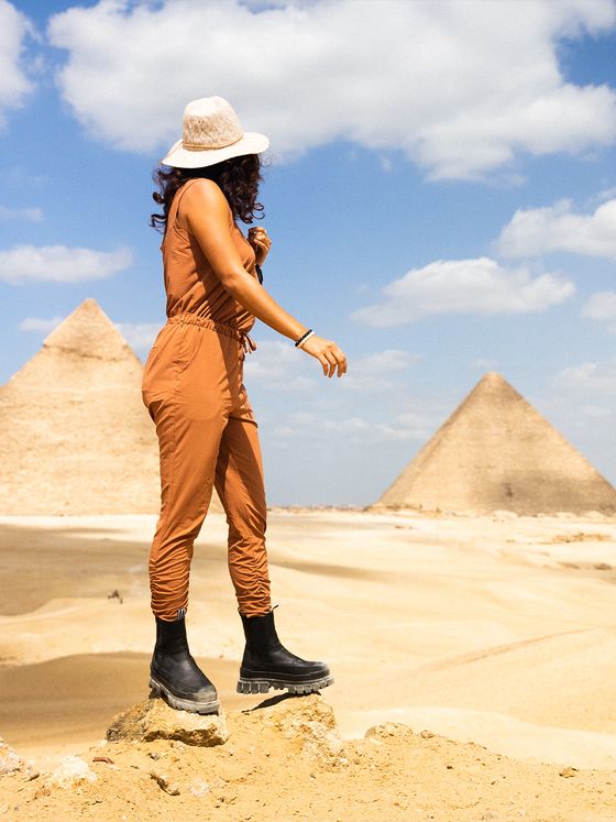 A person in a brown jumpsuit and hat stands on a rock near the pyramids under a blue sky with scattered clouds.