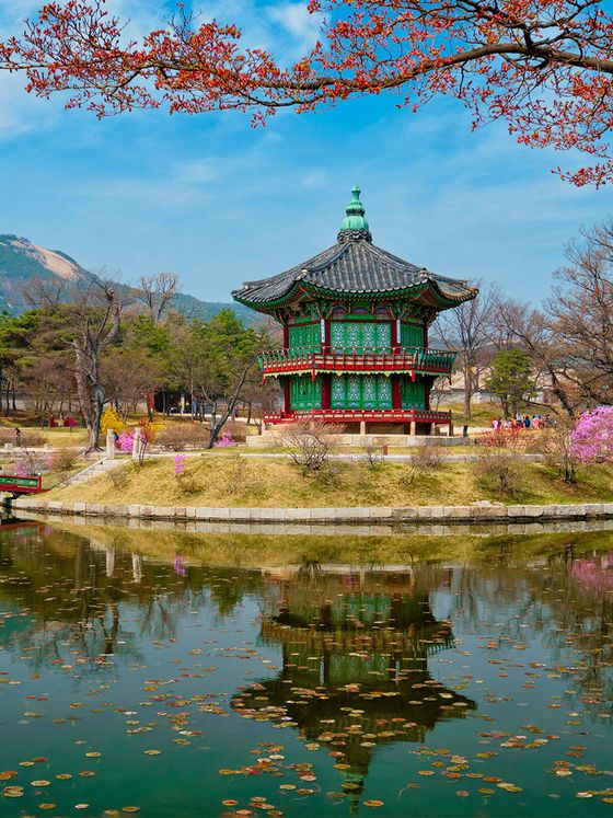 A traditional Korean pavilion by a tranquil pond, surrounded by spring blossoms and trees, with mountains in the background under a blue sky.