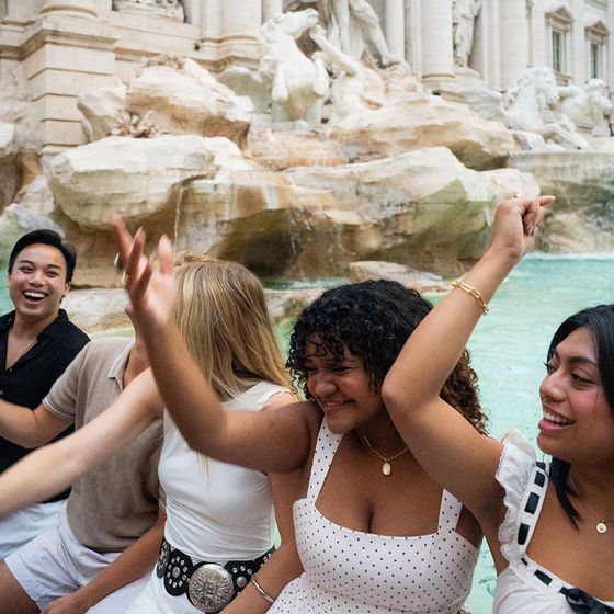 A group of people smiling and raising their arms, sitting by the Trevi Fountain in Rome, with intricate stone sculptures in the background.