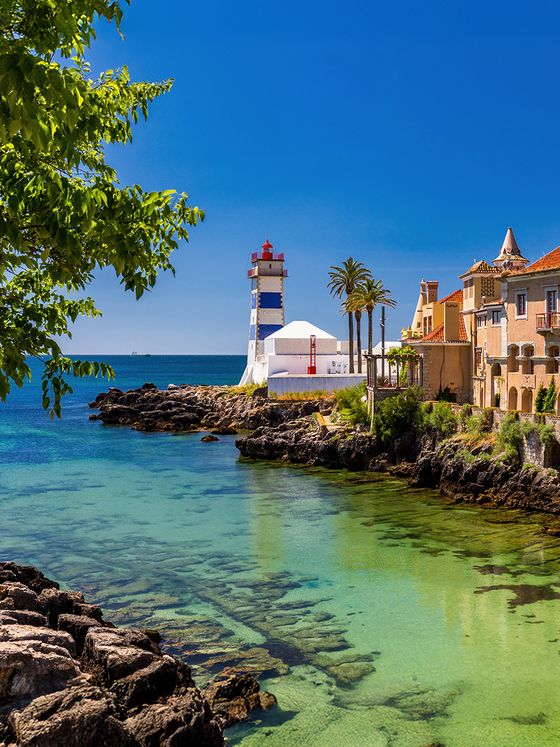 Scenic coastal view featuring a striped lighthouse, clear turquoise waters, rocky shore, palm trees, and historic buildings under a bright blue sky.