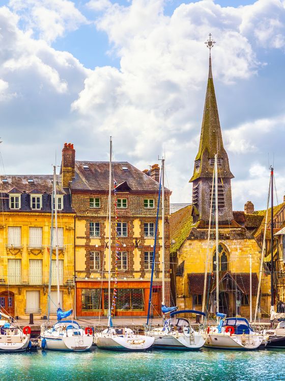A picturesque harbor scene with sailboats docked in front of historic buildings and a tall church steeple under a partly cloudy sky.