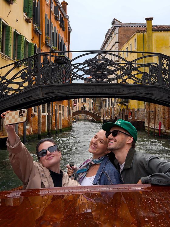 Three people smile and take a selfie on a boat in a Venetian canal, with a decorative bridge and colorful buildings in the background.