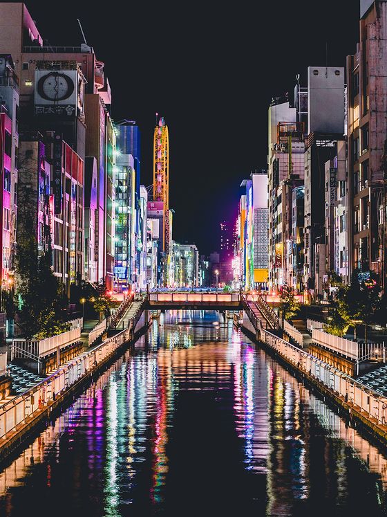 Vibrant cityscape at night with colorful illuminated buildings reflecting in a central canal, lined with railings and streetlights.