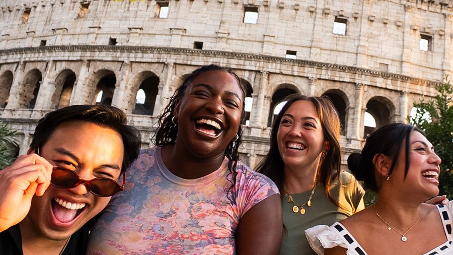 Four people laughing together in front of the Colosseum, with a clear sky and a tree branch overhead.