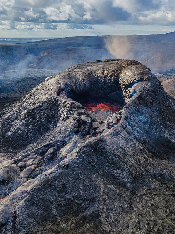 Aerial view of a volcanic crater with glowing lava and surrounding barren landscape under a cloudy sky.