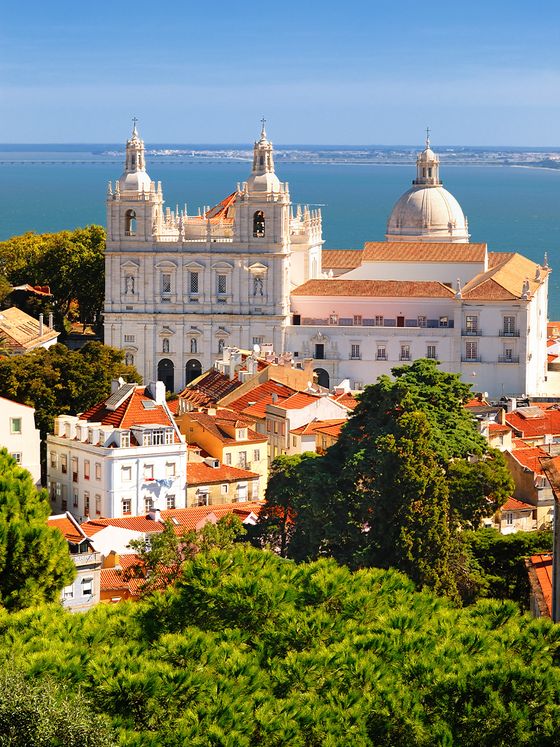Aerial view of São Vicente de Fora Church in Lisbon, with its two bell towers, dome, surrounding red-roofed buildings, and the Tagus River in the background.