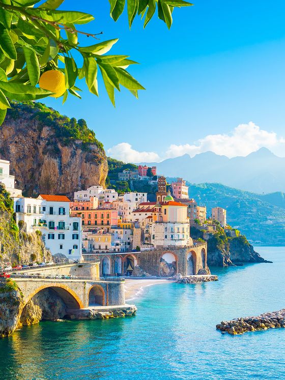 Coastal town with colorful buildings on cliffs above a bright blue sea, with lemon tree branches in the foreground under a clear sky.