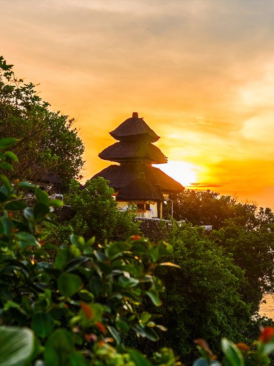 A tiered pagoda-style roof surrounded by lush greenery at sunset, with an orange sky and the sun partially visible on the horizon.