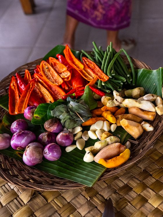 A woven basket with sliced red peppers, green beans, purple onions, garlic cloves, turmeric, and ginger, arranged on banana leaves.