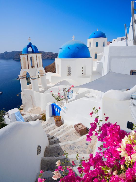 White buildings with blue domes overlook the sea in Santorini, Greece. Stone steps and pink flowers add charm under a clear blue sky.